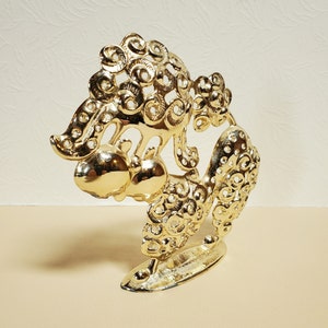Op de afbeelding: Een gouden metalen sculptuur van een gestileerde boom met twee ronde vruchten. De boom is gemaakt van ingewikkelde, draaiende patronen.