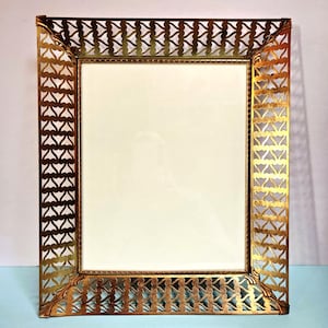 May include: A rectangular gold-colored picture frame with an ornate, openwork design. The frame's border features a repeating pattern of stylized shapes. The interior of the frame is a cream color, ready for a photo or artwork.
