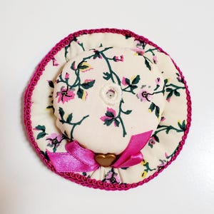 May include: A miniature floral hat with a pink ribbon and trim. The hat has a cream background with pink flowers and green leaves. A gold heart-shaped embellishment is on the pink ribbon.
