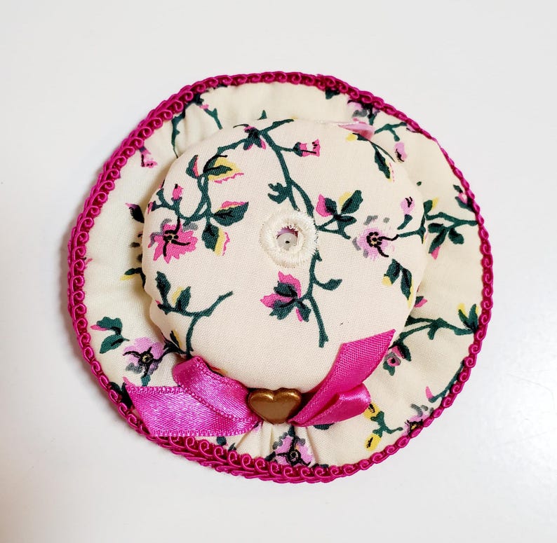 May include: A miniature floral hat with a pink ribbon and trim. The hat has a cream background with pink flowers and green leaves. A gold heart-shaped embellishment is on the pink ribbon.