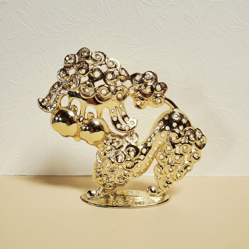 Op de afbeelding: Een gouden metalen sculptuur van een gestileerde draak met ingewikkelde details. De draak is opgerold met zijn kop omhoog en zijn staart om zijn lichaam gewikkeld. Het beeld staat op een kleine voet.