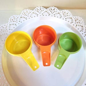 May include: Three measuring cups in yellow, orange, and green. The cups are made of plastic and have a 1/2 cup measurement printed on the yellow cup.