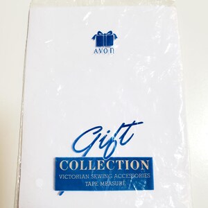 May include: A white packet with blue text and a blue gift box logo. The text reads "AVON" above the gift box. Below, the word "Gift" is written in cursive, and a blue rectangle reads "COLLECTION VICTORIAN SEWING ACCESSORIES TAPE MEASURE".