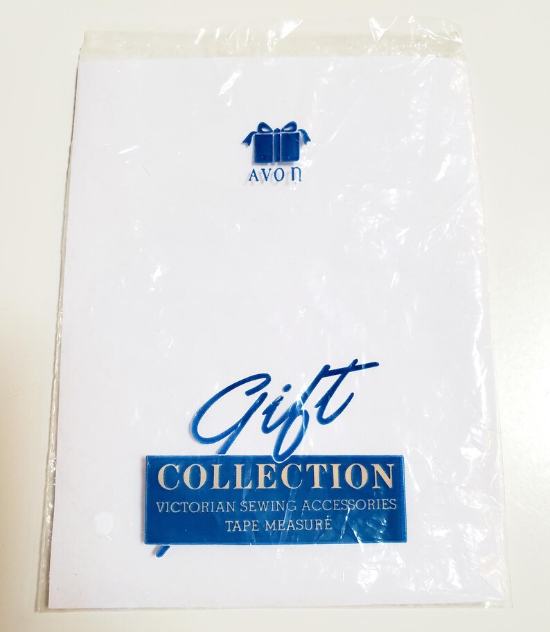 May include: A white packet with blue text and a blue gift box logo. The text reads "AVON" above the gift box. Below, the word "Gift" is written in cursive, and a blue rectangle reads "COLLECTION VICTORIAN SEWING ACCESSORIES TAPE MEASURE".