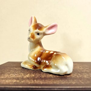 May include: A ceramic figurine of a deer in a resting pose. The deer has brown and white fur with white spots, pink ears, and a white underbelly. The figurine is on a dark brown surface.