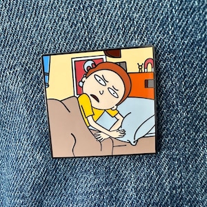 May include: A square enamel pin featuring a cartoon character with brown hair and a yellow shirt lying in bed under a brown blanket. The character has a concerned expression and is resting on a light blue pillow. The background shows a bedroom wall.