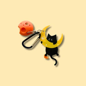 以下が含まれることがあります： 黄色い三日月の上に黒猫がちょこんと座っている、気まぐれなキーホルダー。穴の開いたオレンジ色の球体と、黒いカラビナクリップが付いています。デザインは遊び心のある漫画風です。