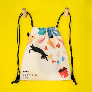 May include: A white drawstring backpack with a colorful design featuring a black cat, climbing holds, and a red backpack. The text "NON VERTICAL LAB" is printed on the front. Black drawstrings hang from wooden pegs.