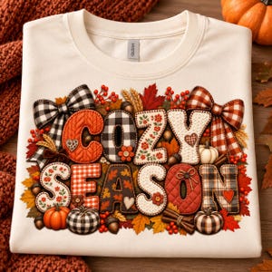May include: A cream-coloured sweatshirt featuring the text COZY SEASON in large, decorative letters. The letters are filled with various patterns including plaid, floral, and knit textures, surrounded by illustrations of pumpkins, leaves, and bows.