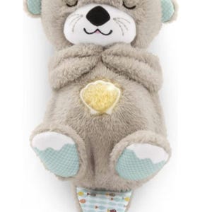 May include: A soft, grey otter-shaped baby toy with closed eyes and a smiling face. It has light blue accents on its ears and feet. A yellow seashell-shaped light is on its chest, and a patterned fabric with fish hangs from its bottom. The text "Any Text" is on the top.