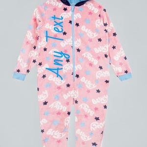 May include: Pink one-piece pyjamas with a character hood. The pyjamas have a zip closure and are patterned with blue stars and the word "Bluey". The hood is navy blue with character details. The text "Any Text" is written on the pyjamas.