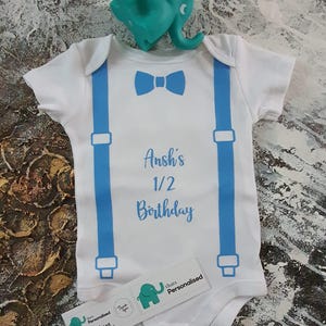 Puede incluir: Body de bebé blanco con tirantes azules y diseño de pajarita. El texto "Ansh's 1/2 Birthday" está impreso en azul. Un juguete de elefante turquesa está colocado encima del body, y una pegatina con un logo de elefante debajo.