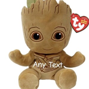 May include: A plush toy of a character with a tree-like appearance, featuring a brown body, large eyes, and small green leaves. The toy has the text "Any Text" and a red heart-shaped tag.