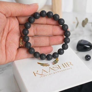 Op de afbeelding: Een armband gemaakt van ronde, poreuze zwarte lavasteen kralen. De armband bevindt zich boven een wit rechthoekig juwelendoosje met de goudkleurige tekst KAASHIR JEWELRY & FRAGRANCE en een gestileerd vleugellogo op het deksel.