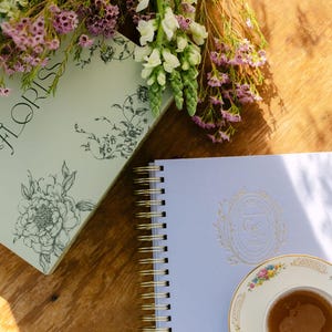 Könnte beinhalten: Ein hellblaues Spiralnotizbuch mit goldenen Akzenten und einem floralen Design, neben einer Teetasse und Untertasse mit floralem Muster. Ein Buch mit dem Wort "FLORISS" und floralen Illustrationen ist ebenfalls vorhanden, zusammen mit Blumen.