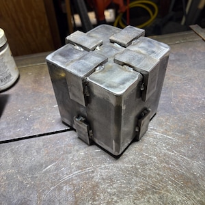 May include: A welded metal cube with rectangular extensions on each face. The cube is made of raw steel, showing weld marks and a metallic finish. The object sits on a metal surface, suggesting a workshop or industrial setting.