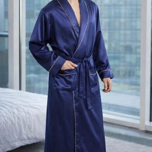 May include: A navy blue satin robe with gold trim. The long robe features a shawl collar, pockets, and a matching belt. The robe is knee-length and appears to be made of a smooth, luxurious fabric. The robe is worn by a person.