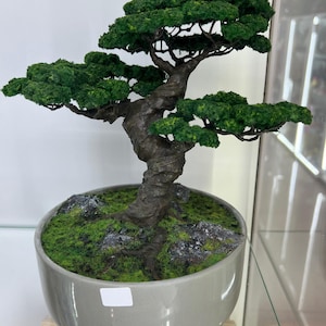 Puede incluir: Un árbol bonsái artificial verde en una maceta de cerámica gris. El árbol tiene un tronco grueso y retorcido y múltiples ramas con follaje verde.