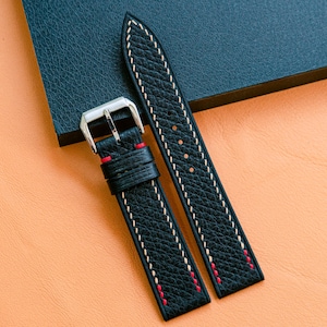 Peut inclure: Bracelet de montre en cuir noir avec coutures blanches et rouges. La sangle a une boucle argentée et est présentée sur une surface orange texturée avec un fond bleu foncé.