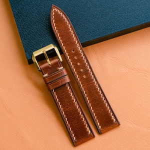 May include: A brown leather watch strap with white stitching and a gold-colored buckle. The strap is displayed on a peach-colored surface with a dark blue leather notebook in the background.