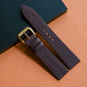 Puede incluir: Una correa de reloj de cuero texturizado en color marrón oscuro con una hebilla de metal dorado y dos trabillas de cuero a juego. La correa presenta un acabado granulado y costuras visibles en los bordes, diseñada para un reloj de pulsera.