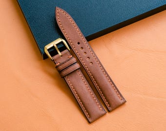 May include: A brown leather watch strap featuring visible stitching along the edges and two leather keepers. The strap includes a polished gold-tone metal buckle with a single prong, designed for secure fastening to a watch case.