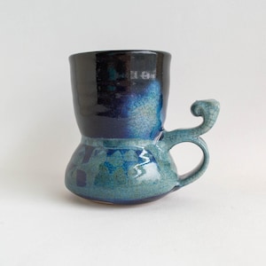 May include: A handmade ceramic mug with a unique shape and a decorative handle. The mug features a gradient of colors, transitioning from black at the top to shades of blue and teal. The handle has a curled design.