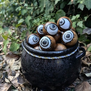 Op de afbeelding: Een zwarte ketel gevuld met houten oogbollen die zijn beschilderd met witte en zwarte details. De ketel staat op een bed van bruine bladeren.