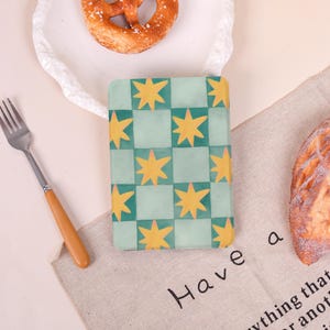 May include: A notebook with a green and yellow star pattern. The notebook is on a beige linen cloth with the words "Have a" printed on it. A pretzel and a fork are also visible. The notebook's design is a grid of green squares with yellow stars.