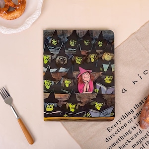 May include: A rectangular notebook featuring a colorful illustration of a classroom filled with green-skinned witches wearing black pointed hats, with one central witch wearing a pink hat and sweater, and the text Black Magic on an open book.