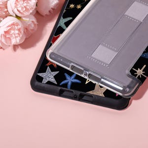 May include: Black phone case with a colorful starfish pattern, topped with a clear protective case. The starfish are in various colors, including blue, silver, and gold. A light gray stand is attached to the clear case.