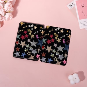 May include: Two black tablet cases with a colorful star pattern. The stars are in various colors, including gold, red, blue, and silver. Some stars have a patterned design. The cases are on a pink surface.