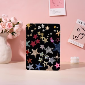 May include: Black tablet case with a colorful star pattern. The stars are in various colors and designs, including gold, silver, red, and blue. The case is rectangular and has rounded corners. The background is a soft pink.