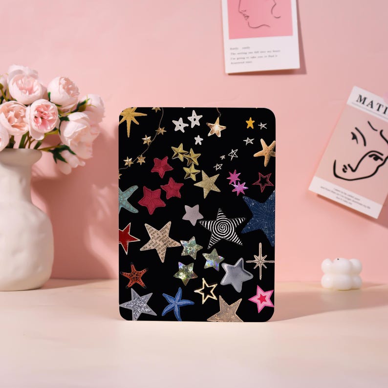 May include: Black tablet case with a colorful star pattern. The stars are in various colors and designs, including gold, silver, red, and blue. The case is rectangular and has rounded corners. The background is a soft pink.