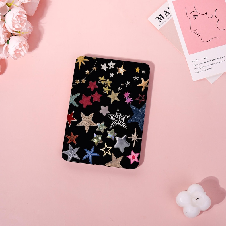 May include: A black rectangular case adorned with a variety of colorful star shapes. The stars are in gold, silver, red, blue, and other colors. Some stars have a glittery finish, while others have a patterned design.