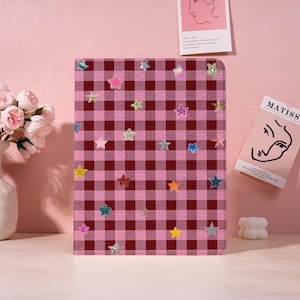 May include: A pink and burgundy checkered notebook cover adorned with various colorful star-shaped embellishments. The notebook is rectangular and stands upright, showcasing a playful and decorative design. The stars are in shades of pink, yellow, blue, and silver.