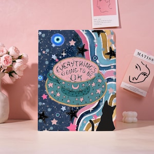 May include: A notebook with a whimsical design featuring a teacup with the text "Everything's Going to Be OK". The cover has a dark blue background with stars, a black cat, and a rainbow. A blue eye symbol is also present.