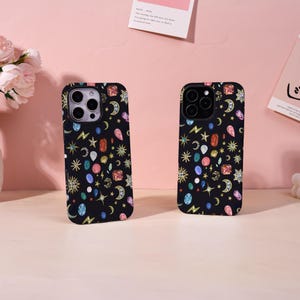May include: Two black smartphone cases featuring a repeating pattern of colorful gemstones, golden crescent moons, and sparkling stars. Each case has a precise cutout for the camera lens array, showcasing a vibrant celestial and jewelry-inspired design.