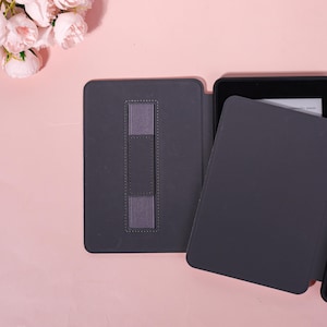 May include: A dark gray e-reader case with a matching elastic hand strap. The case is open, revealing the screen of the e-reader. The background is a light pink surface with a cluster of pink flowers.