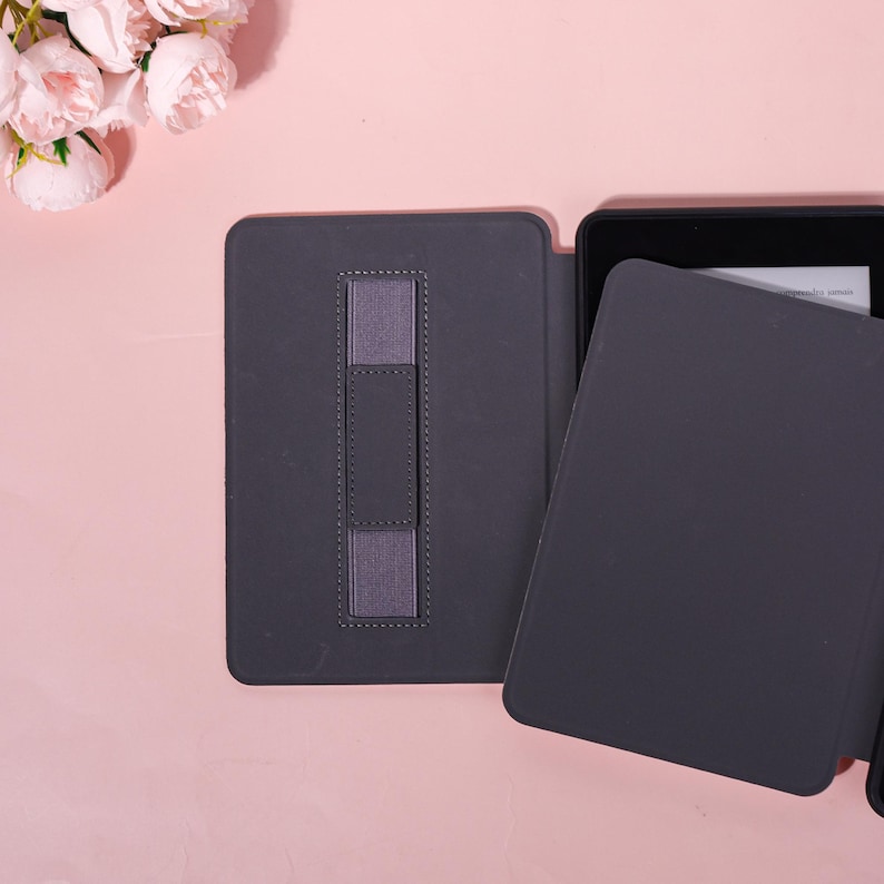 May include: A dark gray e-reader case with a matching elastic hand strap. The case is open, revealing the screen of the e-reader. The background is a light pink surface with a cluster of pink flowers.
