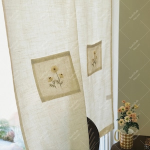 May include: Off-white linen curtain panels with embroidered yellow daisy floral designs. The panels have a square patch with the daisy embroidery. The curtain is hanging in front of a window.