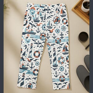 Op de afbeelding: Witte broek met nautisch thema met een herhalend patroon van zeilboten, ankers, vuurtorens, zeemeeuwen en reddingsboeien in blauw, oranje en blauwgroen. De broek is gemaakt van een zacht, comfortabel materiaal.