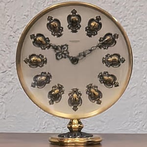 May include: A vintage-style, round clock with a gold-tone frame and base. The clock face is off-white with ornate, dark accents marking the hours. The hands are a dark, decorative metal. The clock is a decorative item.