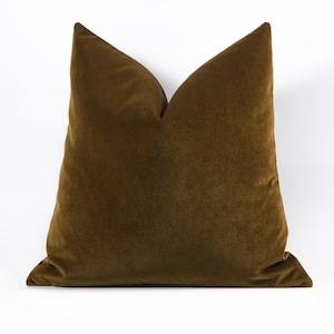 May include: A square throw pillow cover in a deep olive green velvet fabric. The pillow features a distinct karate chop indentation at the top center, creating two pointed corners. The surface has a soft, uniform texture with a subtle sheen.