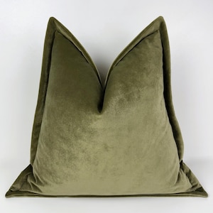 May include: Olive green velvet cushion with a contrasting welt. The square cushion has a soft, plush appearance and a tailored edge. The cushion is a decorative accent for a sofa or bed.