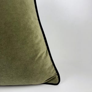 May include: Olive green velvet pillow with a black trim. The pillow has a soft, plush texture and a simple, elegant design. The corner of the pillow is visible against a white background.