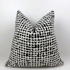 May include: A square decorative pillow with a black and white pattern. The pattern consists of small, irregular black shapes on a white background, creating a textured appearance. The pillow is likely for home decor.