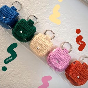 May include: Five crocheted AirPod cases in different colors, hanging on a white wall with a clear acrylic shelf. The colors are blue, green, cream, pink, and orange.