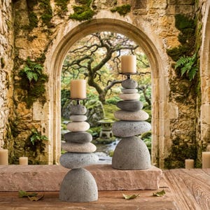 May include: Two candle holders constructed from stacked smooth, rounded river stones in shades of gray and beige. Each holder features a flat metal plate at the top supporting a single cylindrical beige pillar candle, creating a balanced tower.