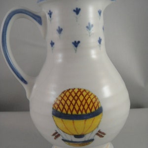 May include: White ceramic pitcher with a blue rim and blue floral pattern. The pitcher features a hand-painted yellow and brown hot air balloon design.
